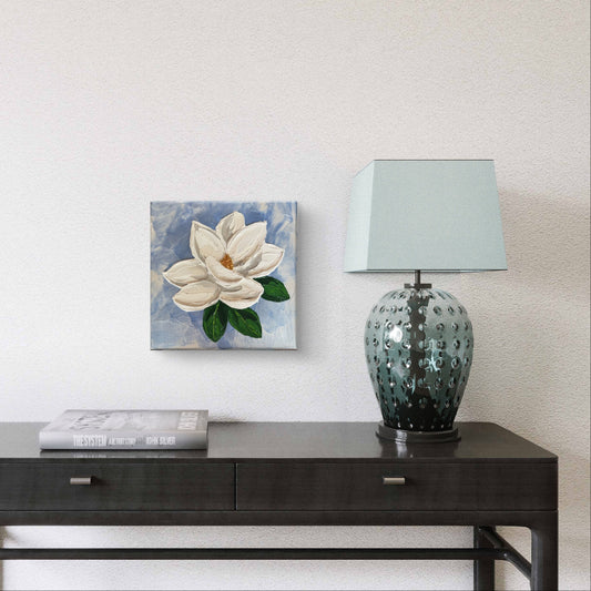 A small square canvas painting of a white magnolia flower with green leaves, displayed on a dark console table next to a blue-green patterned table lamp with a sage green shade.